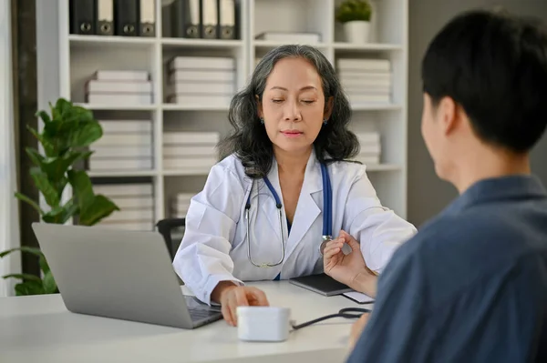 一位专业的亚洲资深女医生正在检查病人的血压和心率 她的桌上有一个监视器在检查室 — 图库照片
