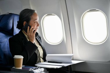 Profesyonel ve başarılı bir Asyalı iş kadını ya da resmi takım elbiseli kadın CEO uçuş sırasında iş ortağıyla telefonda konuşuyor. İş gezisi, havayolu, yolculuk