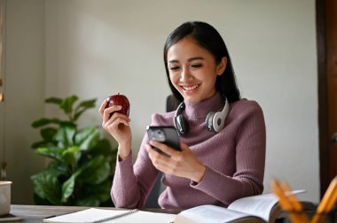 Güzel bir Asyalı kadın evden uzakta çalışırken akıllı telefonuyla sosyal medyada geziniyor..