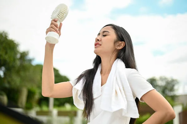 A sweaty Asian woman in sportswear using a portable handy fan, feeling hot and tired after a long run at the park on a sunny day. Sport and lifestyle concept