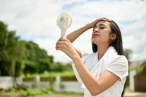 A sweaty Asian woman in sportswear using a portable handy fan, feeling hot and tired after a long run at the park on a sunny day.
