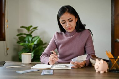 Ciddi bir Asyalı kadın hesap makinesi kullanarak masada oturuyor, elektrik faturalarını kontrol ediyor ve ev giderlerini planlıyor. Tasarruf, bütçe, gelir, giderler, mali 