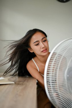 Yorgun ve aşırı ısınmış Asyalı bir kadının portresi sıcak bir yaz gününde oturma odasında dinlenirken kendini elektrikli vantilatörle serinletir..