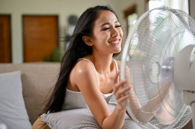 Mutlu ve güzel bir Asyalı kadın sıcak bir yaz gününde oturma odasındaki kanepede dinlenirken elektrikli vantilatörden temiz hava soluyor..
