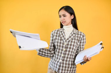 Mutsuz ve kafası karışmış Asyalı genç bir iş kadını elinde belgelerle izole edilmiş sarı bir arka plana karşı duruyor. tatminsiz, sorun, sorun, iş yerinde bunalmış