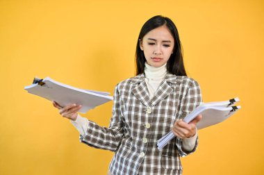 Kafası karışmış ve kafası karışmış genç bir Asyalı iş kadını, izole edilmiş sarı arka planda dururken elindeki belgelere bakıyor..