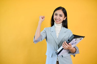 Güzel ve gülümseyen Asyalı genç bir iş kadını iş belgelerini tutuyor ve izole edilmiş sarı arka planda dururken yumruğunu sıkıyor. Başarı, neşelenin, takım çalışması