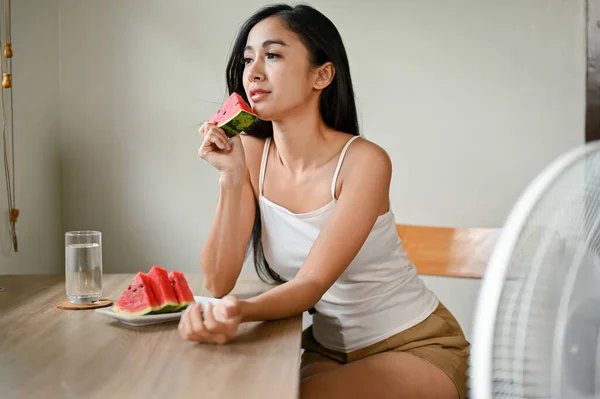 アジアの美しい女性は 暑さと疲れを感じ スイカのスライドを食べて 家で暑い夏の日に自分自身を冷やすために電気ファンの前に座っています — ストック写真