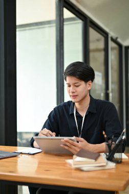 Yakışıklı, Asyalı genç bir ofis çalışanı tabletine odaklanıyor, masasındaki görevleri üzerinde çalışıyor..