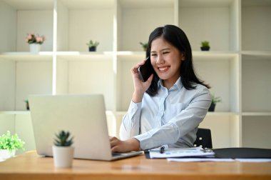 Çekici ve çekici bir Asyalı iş kadını masasında oturmuş iş ortağıyla telefonda iyi bir konuşma yapıyor..