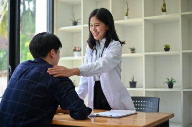 Profesyonel bir Asyalı kadın doktor, hastanedeki sağlık kontrolü sırasında hastanın omzuna dokunarak rahatlık ve güvence sağlar..