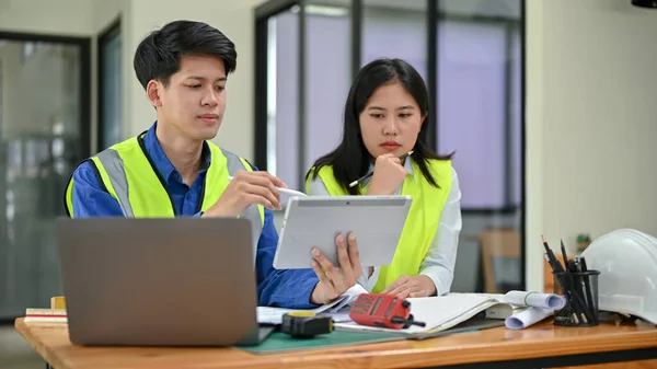 安全ベストを身に着けている2人の専門家と深刻なアジアのエンジニアは タブレット画面を見て 仕事について話し合い オフィスで一緒にプロジェクトに取り組んでいます 工場又は建設技術者 — ストック写真