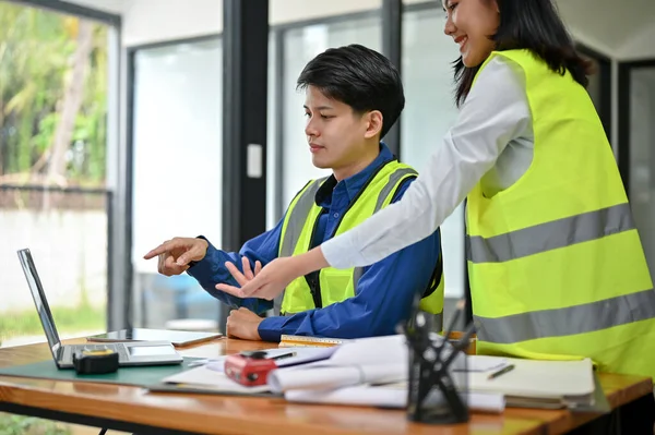 安全性のベストのスマートかつプロフェッショナルなアジアの男性エンジニアは オフィスの女性同僚とデスクトップ上の建物の青写真を議論しています 建設技術者の概念 — ストック写真