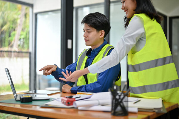 A smart and professional Asian male engineer in a safety vest is discussing a building blueprint on a desktop with his female colleague in the office. Construction engineer concept