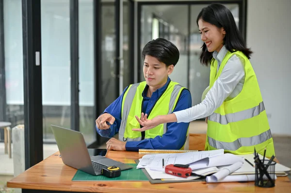 安全性のベストのスマートでプロフェッショナルなアジアの男性エンジニアは ラップトップ画面を見て オフィスの彼の女性同僚とデスクトップ上の建物の青写真を議論しています — ストック写真