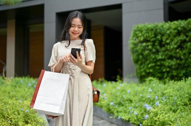 A beautiful Asian woman in a beautiful minimal dress is texting someone on her smartphone while walking on the street with her shopping bags. Urban lifestyle concept