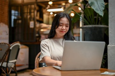 Güzel genç bir Asyalı kadın güzel bir kahve dükkanında otururken bilgisayarını iş işlerini yönetmek için kullanıyor. Uzak çalışma ve kentsel yaşam tarzı kavramı