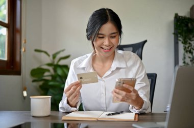 Genç ve güzel Asyalı bir kadın elinde akıllı telefon ve kredi kartıyla online alışveriş yapıyor.. 