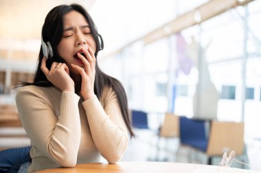 Yorgun ve uykulu genç bir Asyalı kadın müzik dinlerken esniyor ve bir kahve dükkanında dinleniyor..