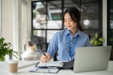 Profesyonel ve güzel bir Asyalı iş kadını evrakları imzalıyor ve masasında, modern bir ofiste çalışıyor..