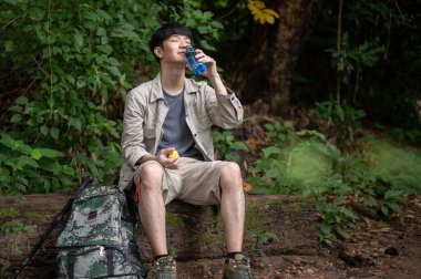 Mutlu ve rahat Asyalı bir erkek yürüyüşçü ormanda yürüyüş yaparken bir kayanın üzerinde oturur, bir şişeden su içer ve dinlenir..