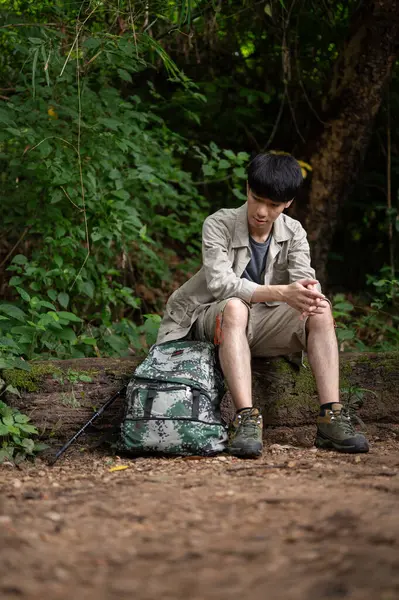 stock image An exhausted young Asian male hiker sits on a stone, resting and taking a break during his hiking trip in the forest alone.