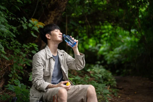 stock image A happy and relaxed Asian male hiker sits on a rock, drinking water, while resting during hiking in the forest.