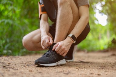 Yeşil ormanda koşarken ayakkabı bağcıklarını bağlayan sportif ve aktif Asyalı genç bir adamın yakın plan görüntüsü..