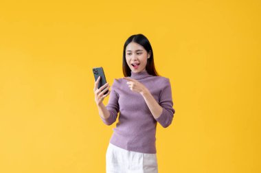 Güzel bir Asyalı kadın akıllı telefon ekranını işaret ederek mutlu ve şaşırtıcı bir yüzle bakıyor. izole edilmiş sarı arkaplan.