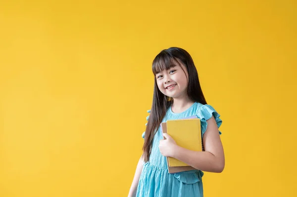 站在孤立的黄色背景下 手里拿着书的可爱而微笑的年轻亚洲女孩或小学生 — 图库照片