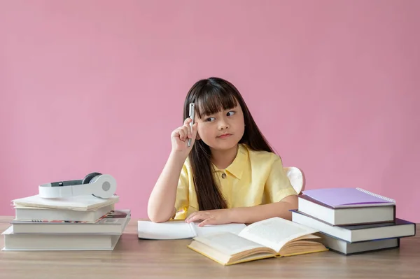 一位可爱的年轻亚洲女孩坐在自己的学习桌上 面对着一个孤立的粉色背景 脸上流露出沉思和思考的表情 小学生概念 — 图库照片