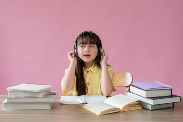 一位可爱的年轻亚洲女孩戴着耳机 在自己的学习桌上用一个孤立的粉色背景学习 小学生概念 — 图库照片