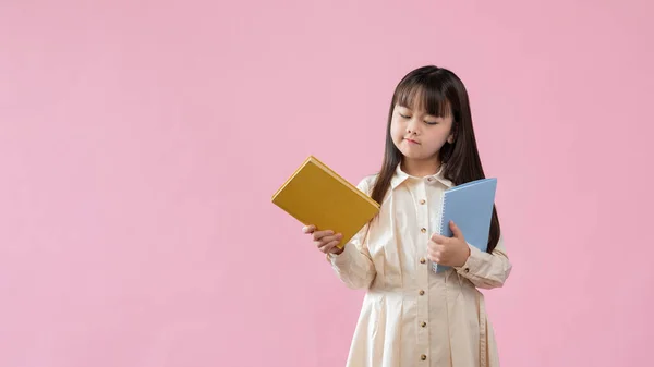 一个体贴可爱的年轻亚洲女孩站在一个孤立的粉色工作室的背景下 用怀疑和沉思的脸看着手中的书 教育和儿童概念 — 图库照片