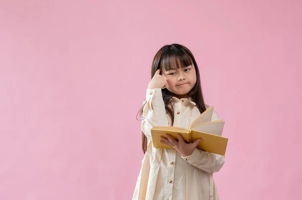一个可爱又体贴的年轻亚洲女孩拿着一本翻开的书 脸上带着沉思和怀疑的表情 站在一个孤立的粉色工作室的背景下 小学生 — 图库照片