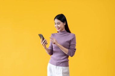 Güzel bir Asyalı kadın akıllı telefonunu kullanıyor, sohbet ediyor, SMS veya e-posta gönderiyor, sosyal medyada kayıyor, blogunu güncelliyor, mobil uygulamalar kullanıyor ve çevrimiçi alışverişin tadını çıkarıyor. izole edilmiş sarı arkaplan