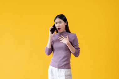 Şok olmuş ve panikleyen Asyalı bir kadın telefonda biriyle konuşuyor, izole edilmiş sarı bir arka plana karşı duruyor. Kötü haber, inanılmaz, korkmuş, korkmuş