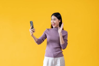 Güzel bir Asyalı kadın elini sallıyor, akıllı telefonuyla arkadaşlarıyla konuşurken selam veriyor, izole edilmiş sarı bir arka plana karşı duruyor..