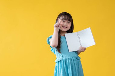 Mavi elbiseli parlak ve güzel bir Asyalı kız boş sayfaları olan açık bir kitap tutuyor, parmağını kaldırıyor, gülümsüyor ve kameraya bakıyor. izole edilmiş sarı arkaplan