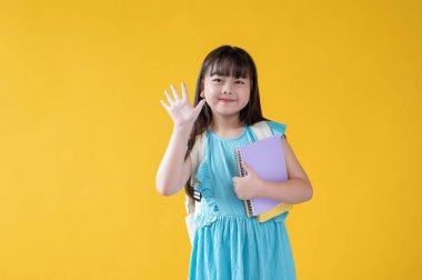 Şirin mavi elbiseli güzel ve arkadaş canlısı bir Asyalı kız elini kaldırıyor ve arkadaşlarına merhaba diyor. İzole edilmiş sarı arka plan. Öğrenci, ilkokul öğrencisi, çocuklar, pozitif çocuk.