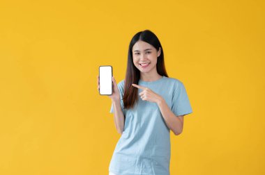 Mavi tişörtlü çekici ve gülümseyen Asyalı bir kadın, izole edilmiş sarı bir arkaplanda dikilirken akıllı telefonunu işaret ediyor. Akıllı telefon beyaz ekran modeli