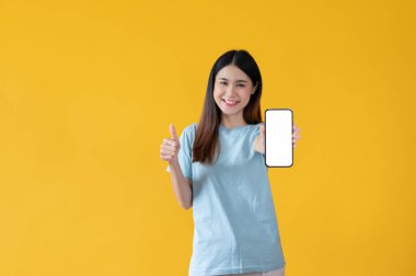 Güzel bir Asyalı kadın baş parmağını kaldırıyor ve kameraya elinde bir akıllı telefonun beyaz ekran ile izole edilmiş sarı arka planda duruyor..