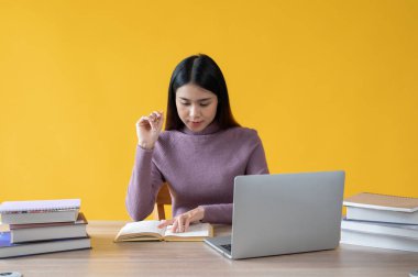 Güzel bir Asyalı kadın masasında izole edilmiş sarı bir arkaplan karşısında kitap okumaya odaklanıyor. İş kadını, üniversite öğrencisi.