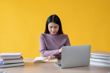 Güzel bir Asyalı kadın bir kitap okumaya odaklanıyor ve masasındaki ödevi üzerinde çalışıyor, izole edilmiş sarı bir arka plana karşı. İş kadını, üniversite öğrencisi.