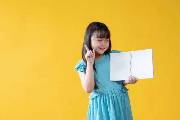 青いドレスの可愛いアジアの少女が 空っぽのページで開いた本を持ち 指を差し出し 笑顔を浮かべ カメラを見ている 隔離された黄色の背景 — ストック写真