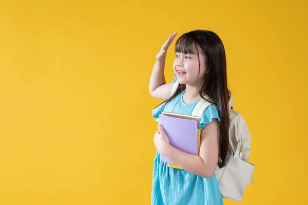 一个穿着漂亮蓝色衣服和背包的积极友善的亚洲小女孩正在向她的朋友们致意 把目光投向一边 挥挥手打招呼 孤立的黄色背景 — 图库照片