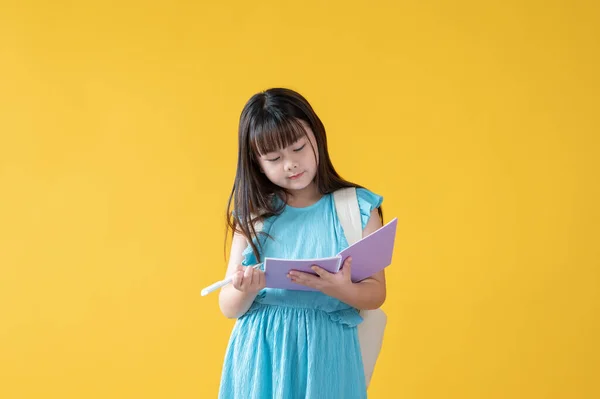 一个可爱的亚洲小女孩站在一个孤立的黄色背景下 在她的书里写着什么或者做作业 小学生 — 图库照片