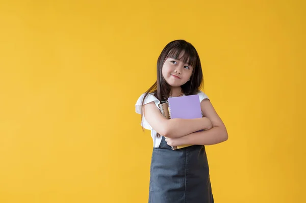 一个穿着漂亮牛仔裤裙的可爱的亚洲小女孩正从旁边看过去 手里拿着书 站在一个孤立的黄色背景下 快乐的女孩 女学生 小学生 — 图库照片