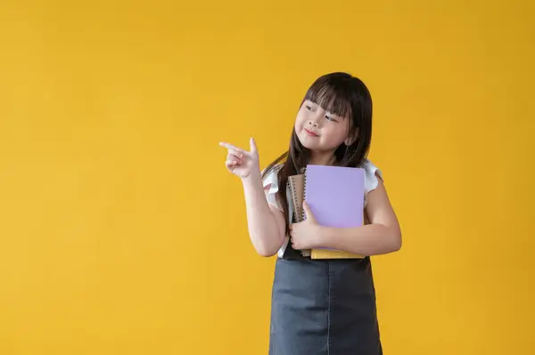 一个穿着漂亮牛仔裤裙的可爱而又积极的亚洲小女孩把手指放在空荡荡的地方 拿着书 站在孤立的黄色背景下 女学生 — 图库照片