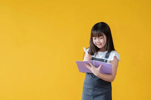 一个快乐而可爱的亚洲小女孩正站在一个孤立的黄色背景下 在她的书里写着什么 小学生 — 图库照片