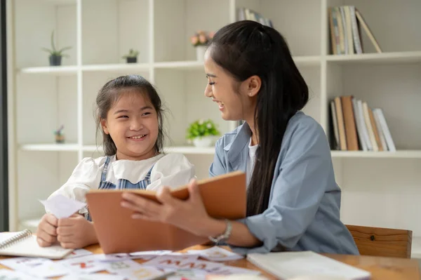 A cheerful Asian female private teacher and a young girl are laughing and enjoying the English class together. Kids education concept
